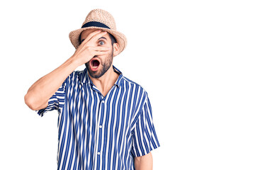 Young handsome man with beard wearing summer hat and striped shirt peeking in shock covering face and eyes with hand, looking through fingers with embarrassed expression.