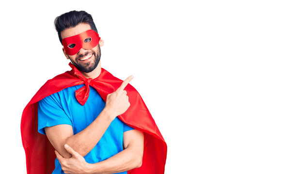 Young handsome man with beard wearing super hero costume with a big smile on face, pointing with hand and finger to the side looking at the camera.