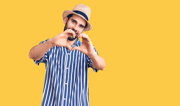 Young handsome man with beard wearing summer hat and striped shirt smiling in love doing heart symbol shape with hands. romantic concept.
