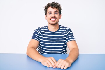 Young caucasian man with curly hair wearing casual clothes sitting on the table looking positive and happy standing and smiling with a confident smile showing teeth