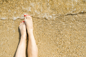 Top view of beautiful female feet with bright red pedicure on the sand of the beach. The sea wave washes women's feet. Relaxation and enjoyment during your seaside holiday. Copy space