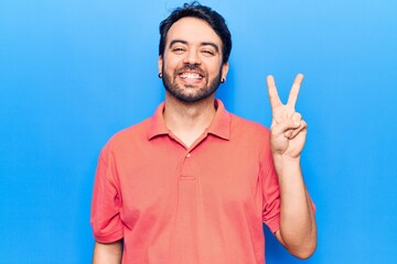 Fototapeta premium Young hispanic man wearing casual clothes smiling with happy face winking at the camera doing victory sign. number two.