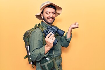 Young hispanic man wearing explorer clothes holding binoculars screaming proud, celebrating victory and success very excited with raised arm