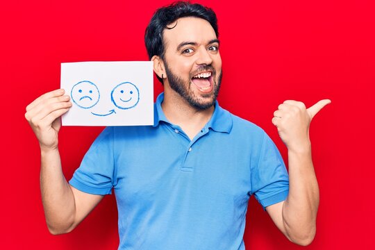 Young hispanic man holding sad to happy emotion paper pointing thumb up to the side smiling happy with open mouth
