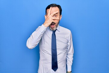 Middle age handsome business man wearing elegant tie standing over isolated blue background peeking in shock covering face and eyes with hand, looking through fingers afraid