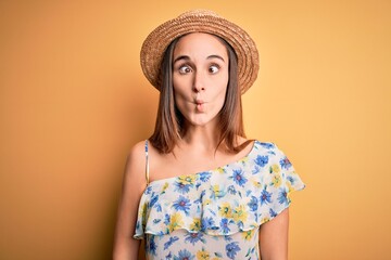 Young beautiful woman wearing casual t-shirt and summer hat over isolated yellow background making fish face with lips, crazy and comical gesture. Funny expression.