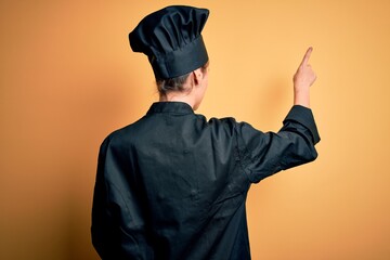 Young beautiful chef woman wearing cooker uniform and hat standing over yellow background Posing backwards pointing ahead with finger hand