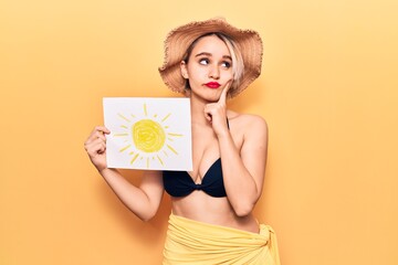 Young beautiful blonde woman wearing bikini and summer hat holding sun draw serious face thinking about question with hand on chin, thoughtful about confusing idea