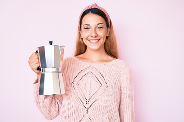 Young brunette woman holding italian coffee maker looking positive and happy standing and smiling...