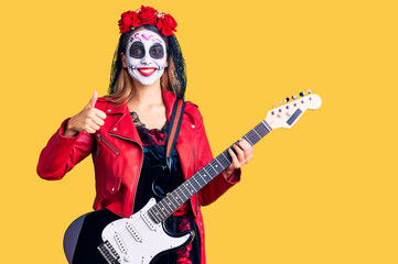 Obraz na płótnie Canvas Woman wearing day of the dead costume playing electric guitar smiling happy and positive, thumb up doing excellent and approval sign