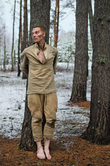 young man in military uniform tied to the trunk of an old tree barefoot in the winter forest