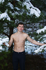young man without a shirt in the winter forest stands under a tree