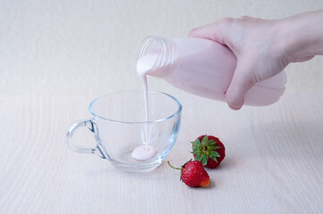 Red strawberries, a woman's hand pours yogurt from a bottle into a glass mug on the table