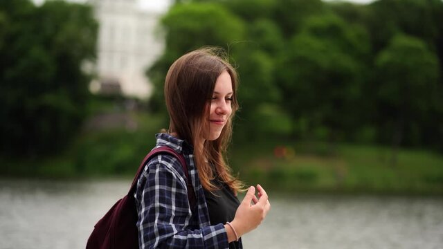 Young woman standing near lake. Side view of young female in checkered shirt smiling and looking away while standing on blurred background of lake and estate
