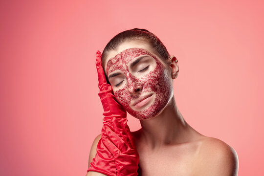 An adorable woman with a strawberry scrub on her face and red gloves touches her face. Natural cosmetic. Pink studio background