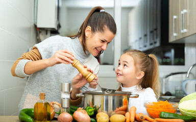 Little daughter helping cooking soup and mother add pepper to pan