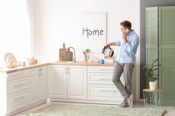 Man with mobile phone pouring hot boiled water from electric kettle into cup at home
