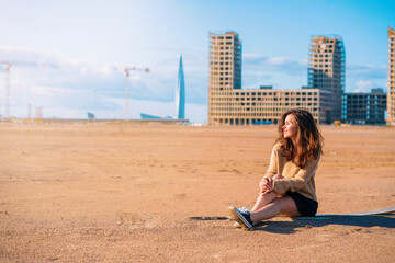 Fototapeta premium A young woman in a cozy sweater and skirt with a backpack sits on the sand, behind her new buildings and skyscrapers under construction