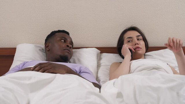 Portrait of interracial couple in bed. Black man asking his white girlfriend to hang up phone. Girl ignoring boyfriend and keeps talking