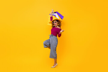 Full body portrait of smiling fashionable young African American woman raising hand with shopping bags in isolated yellow background for summer sale concept