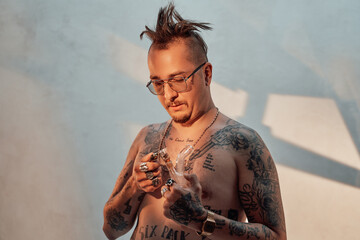 Shirtless man with tattooed body and freaky haircut holds the bong with cannabis. Cannabis...