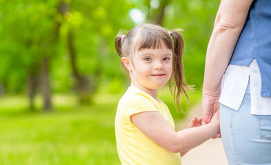 Obraz premium Happy little girl with syndrom down holds mom's hand in a sunny summer park. Empty space for text