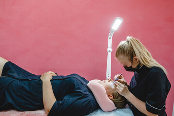 Blonde master in black uniform in the process of eyelash extension in the beauty salon