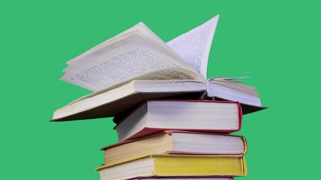 One interesting open book with flying leaves lies on a stack of books with colored covers, waiting for its readers. Studio shot on green background. Reading, leisure, self-development concept.