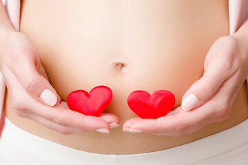 Young woman hands holding two red bright hearts on naked belly. Emotional loving moment in pregnancy time. Twins babies expectation. Love, happiness and safety concept. Closeup. Front view.