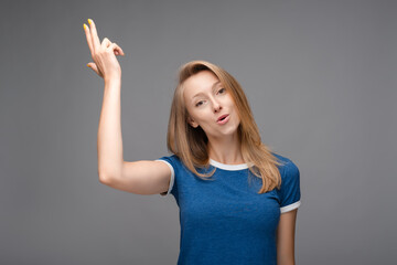 Funny young blonde female shoots in temple, tilts head, dressed in casual blue t shirt, demonstrates suicide gesture, isolated on gray background