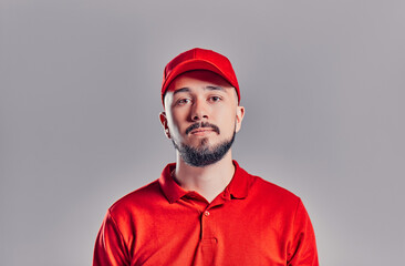 Bearded young delivery man in red t-shirt and cap isolated on gray background. Home delivery service.