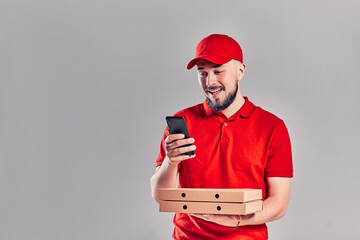 Bearded young delivery man in red T-shirt and cap with pizza boxes and with smartphone isolated on gray background. Fast home delivery.