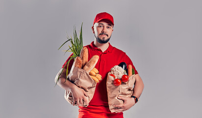 Young bearded delivery man in red uniform holds two large heavy packages with bread and vegetables isolated on gray background.