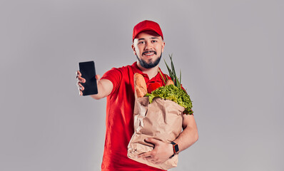 Young bearded delivery man in red uniform holds package with bread and vegetables and shows smartphone screen isolated on gray background.