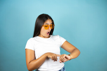 Young beautiful woman wearing sunglasses  standing over isolated blue background surprised, looking...