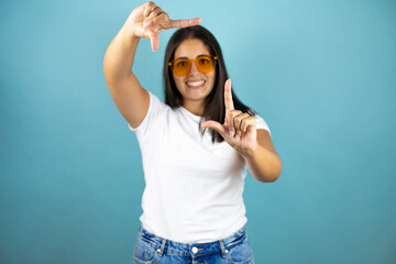 Young beautiful woman wearing sunglasses standing over isolated blue background smiling making frame with hands and fingers with happy face