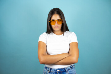 Young beautiful woman wearing sunglasses over blue background skeptic and nervous, disapproving expression on face with crossed arms