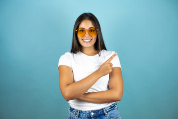 Young beautiful woman wearing sunglasses standing over isolated blue background smiling happy pointing with hand and finger to the side