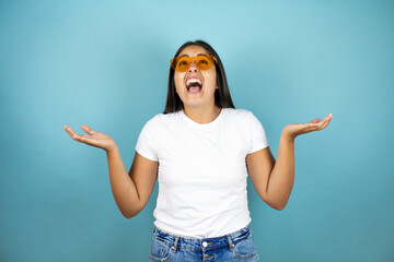 Young beautiful woman wearing sunglasses over isolated blue background clueless and happy...