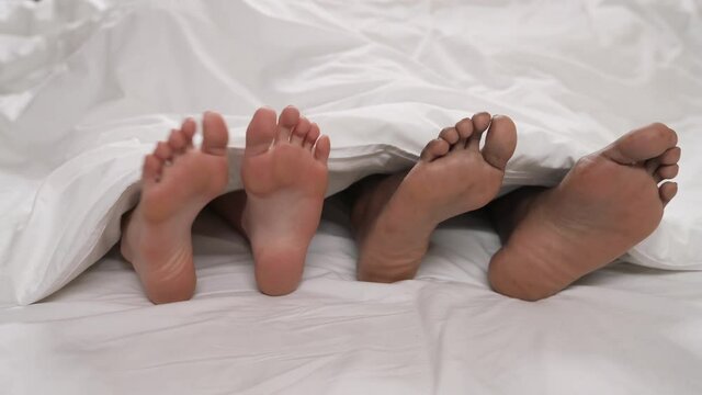 Close-up of swinging side to side bare feet of African man and Caucasian woman lying under white blanket. Love and family concept