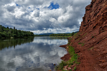 A natural landscape with clouds reflecting on the surface of a calm river with green forest and red soil cliff