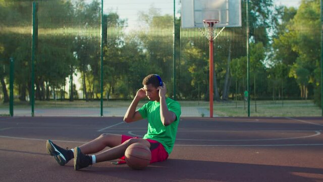 Positive Handsome Street Basketball Player Sitting On Outdoor Court, Wearing Wireless Headphones And Listening To Music Using Cellphone Mp3 Player In Early Morning While Relaxing After Basketball Game