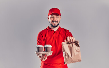 Bearded young man in red T-shirt and cap holds package with fast food and coffee isolated on gray background. Fast delivery from the restaurant to your home.