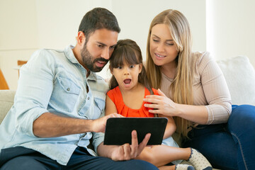 Positive parents and excited little daughter sitting on couch, using tablet, watching footages or movie. Front view. Internet or family leisure time concept