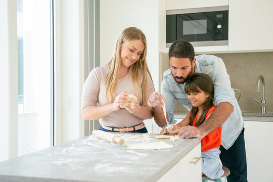 Happy mom and dad teaching daughter to knead dough at kitchen table with flour powder. Young couple and their girl baking together. Family cooking concept
