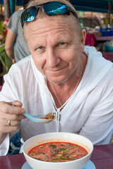 adult elderly man in a white tunic eats a spicy Thai soup Tom Yam in a cafe. Gastronomic tourism in Asia. Spicy traditional food.