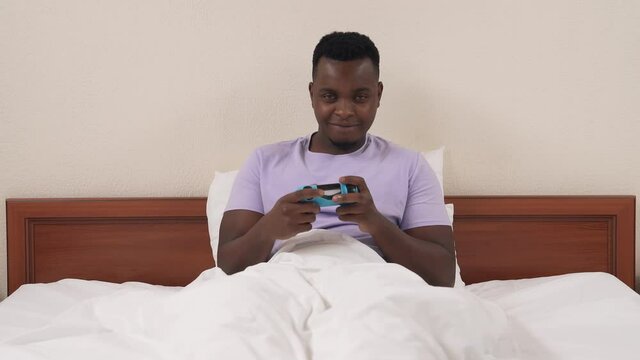 Portrait of black man lying in bed and playing console. Caucasian woman's legs appear in front of camera and taking off lace panties. Astonished guy throws joystick away