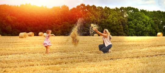 Naklejka na ściany i meble Banner with mom and daughter caucasians together on a mown wheat field throw hay where there are huge sheaves. Family fun and fun. Nature. Harvesting. Agriculture. Travel and vacation concept
