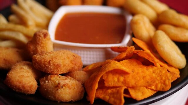 Slow motion closeup of a platter of fries, nachos, wedges, and fish fingers on a black plate with a cup of dipping sauce in the middle