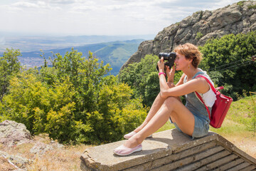 The girl takes pictures with a camera of summer landscapes from a high mountain. Seasonal travel and hiking. Beautiful landscape with rocks and forest and a blue valley.